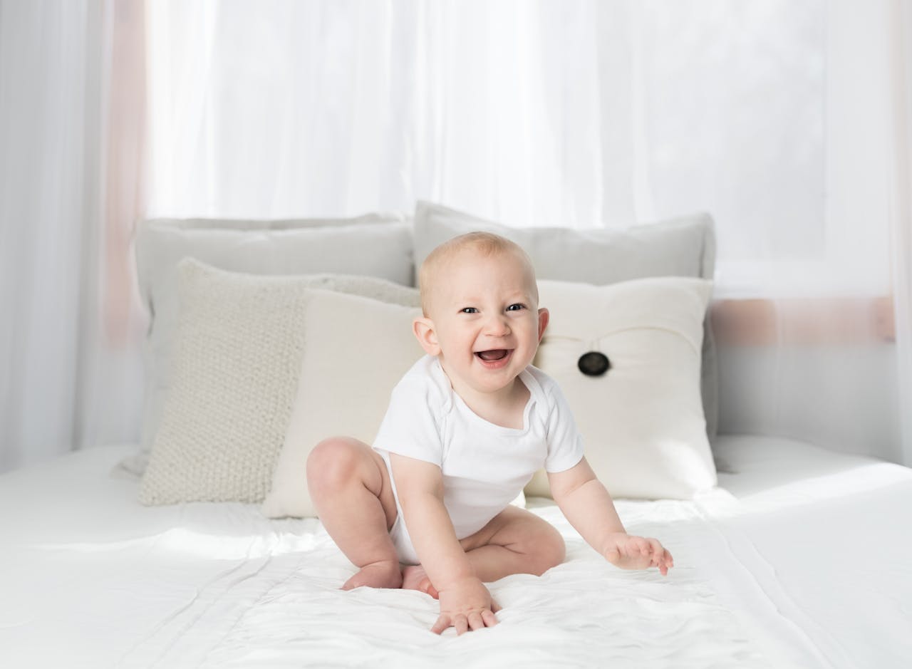Cute baby sitting and smiling on a white bed in a bright, airy bedroom.